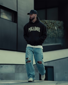 A person is walking outdoors, dressed in a black hoodie with 'PEGADOR' written on it, a black cap, ripped jeans, and white sneakers. The background includes modern architecture with concrete and glass elements, and some greenery is visible.
