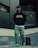 A person is walking outdoors, dressed in a black hoodie with 'PEGADOR' written on it, a black cap, ripped jeans, and white sneakers. The background includes modern architecture with concrete and glass elements, and some greenery is visible.