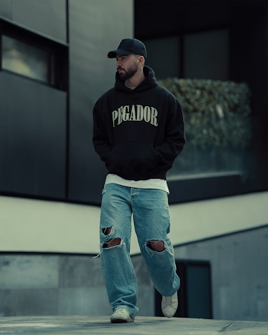 A person is walking outdoors, dressed in a black hoodie with 'PEGADOR' written on it, a black cap, ripped jeans, and white sneakers. The background includes modern architecture with concrete and glass elements, and some greenery is visible.