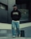 A person is walking outdoors, dressed in a black hoodie with 'PEGADOR' written on it, a black cap, ripped jeans, and white sneakers. The background includes modern architecture with concrete and glass elements, and some greenery is visible.