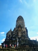 A historic stone temple structure with an ornate spire, surrounded by several tourists who are climbing the steps and exploring the site. The temple is set against a clear blue sky, indicating a bright, sunny day.