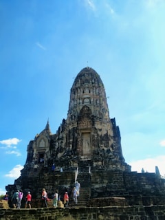 A historic stone temple structure with an ornate spire, surrounded by several tourists who are climbing the steps and exploring the site. The temple is set against a clear blue sky, indicating a bright, sunny day.