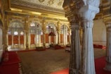 An intricately decorated interior featuring tall, ornate pillars and arched doorways adorned with elaborate carvings and colorful stained glass. The space is furnished with rich red carpets and cushions, contributing to a luxurious and historical ambiance. The walls and ceiling are elaborately designed with detailed patterns and motifs, showcasing a lavish and regal architecture.