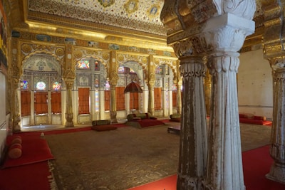 An intricately decorated interior featuring tall, ornate pillars and arched doorways adorned with elaborate carvings and colorful stained glass. The space is furnished with rich red carpets and cushions, contributing to a luxurious and historical ambiance. The walls and ceiling are elaborately designed with detailed patterns and motifs, showcasing a lavish and regal architecture.