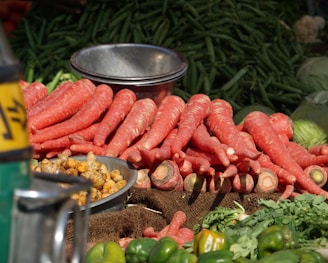 A pile of large, fresh reddish-orange carrots is prominently displayed on a market stall. Next to the carrots, there is a metal bowl containing small, brownish root vegetables. In the background, a heap of green chili peppers and some cabbage are visible. The setting is outdoors, and the produce is arranged neatly on burlap sacks, suggesting a local market or farmer’s stall.