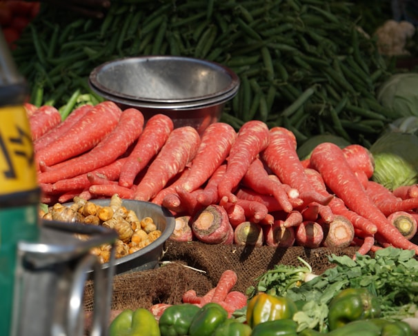 A pile of large, fresh reddish-orange carrots is prominently displayed on a market stall. Next to the carrots, there is a metal bowl containing small, brownish root vegetables. In the background, a heap of green chili peppers and some cabbage are visible. The setting is outdoors, and the produce is arranged neatly on burlap sacks, suggesting a local market or farmer’s stall.