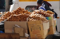 Stacks of rusks and bread snacks fill large cardboard boxes, placed on a street vendor cart. A person stands nearby, partially visible, with the backdrop of a parked vehicle.