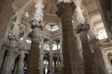 An intricately carved marble interior featuring ornately detailed columns and arches, showcasing magnificent craftsmanship. The structure has a vast and airy feel with soft, diffused lighting entering from above.