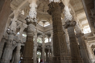 An intricately carved marble interior featuring ornately detailed columns and arches, showcasing magnificent craftsmanship. The structure has a vast and airy feel with soft, diffused lighting entering from above.