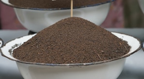 A large mound of fine, dark brown granules is piled high on a round, white enamel bowl with a blue rim. The texture appears coarse and the granules vary slightly in size. The background is softly out of focus, highlighting the piled substance as the main focal point of the image.