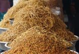 A market stall displaying large piles of various crunchy snacks and foods in metal trays. The foreground features a colorful mixture of small, round pieces and other crispy items. Behind it is a heap of thin, golden-brown noodles.