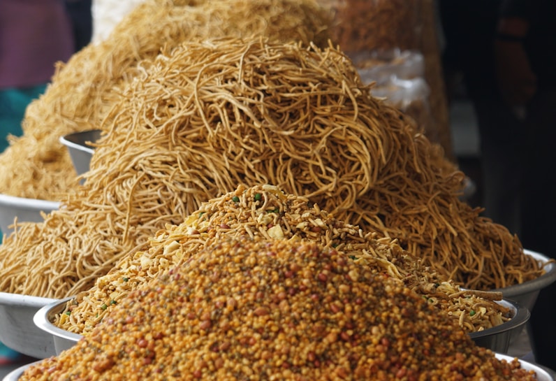 A market stall displaying large piles of various crunchy snacks and foods in metal trays. The foreground features a colorful mixture of small, round pieces and other crispy items. Behind it is a heap of thin, golden-brown noodles.