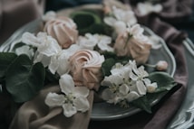 A decorative arrangement featuring delicate white flowers and leaves artfully placed on a soft-colored plate. The composition includes intricately folded fabric, contributing to a serene and elegant presentation.
