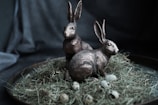 Two rabbit sculptures are positioned on a bed of dried grass, surrounded by quail eggs. The setting appears to be on a round tray with a dark, textured background.