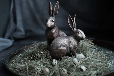 Two rabbit sculptures are positioned on a bed of dried grass, surrounded by quail eggs. The setting appears to be on a round tray with a dark, textured background.