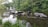 Stones form a rustic path across a tranquil pond surrounded by lush green trees. In the distance, fountains add a gentle dynamic to the serene setting. The water reflects the greenery and overcast sky, creating a peaceful landscape.