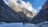 A group of people is walking on a frozen riverbed surrounded by towering rocky cliffs. The landscape features rugged mountains rising majestically under a partly cloudy sky with patches of blue.