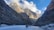 A group of people is walking on a frozen riverbed surrounded by towering rocky cliffs. The landscape features rugged mountains rising majestically under a partly cloudy sky with patches of blue.