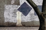A textured concrete wall features abstract shapes including a black square and unpainted gaps revealing a brick pattern underneath. In the foreground, a large tree trunk extends diagonally, contrasting with the wall. The ground has scattered debris and small twigs.