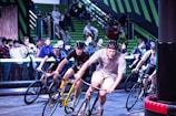 A group of cyclists competes in an indoor race, navigating a track with intense focus. They wear helmets and sport attire, with a crowd of spectators watching intently from the green bleachers in the background.