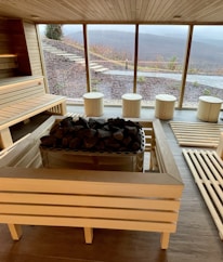 A spacious sauna room with wooden benches and a central stone heater. Large windows reveal an outdoor view of a hilly landscape, with steps leading up a slope. The interior has a bright, clean look with natural wood paneling and flooring.