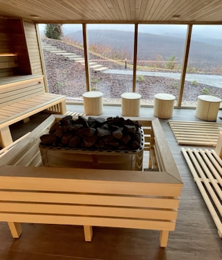 A spacious sauna room with wooden benches and a central stone heater. Large windows reveal an outdoor view of a hilly landscape, with steps leading up a slope. The interior has a bright, clean look with natural wood paneling and flooring.