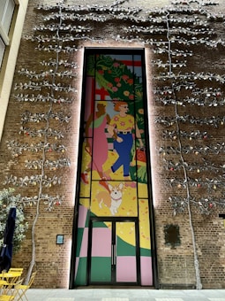 A vibrant mural on a brick wall depicts two people walking a dog. The artwork is colorful, showing one person wearing a pink dress and the other in a shirt with flowers and blue pants. Large green plants and abstract shapes fill the background. The wall is also adorned with metal vines and small lights.