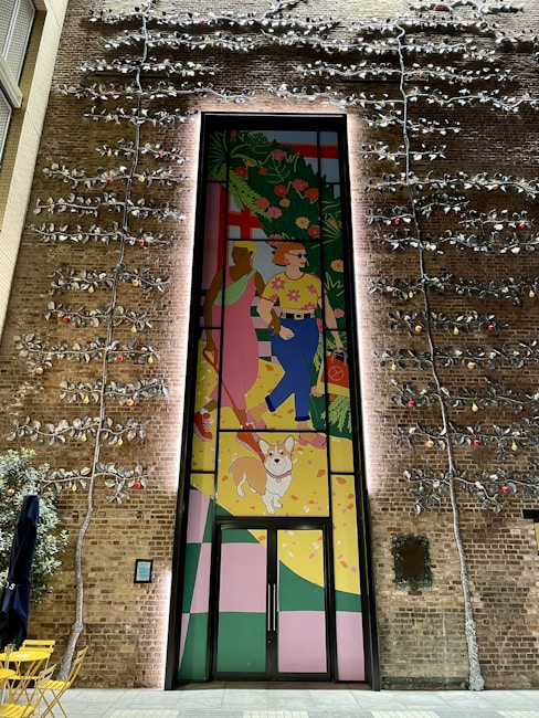 A vibrant mural on a brick wall depicts two people walking a dog. The artwork is colorful, showing one person wearing a pink dress and the other in a shirt with flowers and blue pants. Large green plants and abstract shapes fill the background. The wall is also adorned with metal vines and small lights.