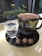 A black coffee cup with the words 'K P Koffee' sits on a black saucer alongside a small glass of sparkling water and three round chocolate truffles. The table surface is glossy, and there is a background view of a window with some green plants visible outside.
