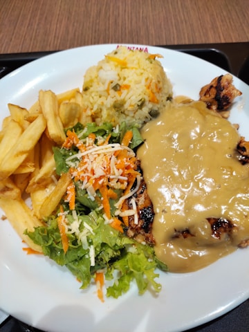 A plate with a serving of seasoned rice, golden fries, fresh salad garnished with shredded carrots and cheese, and grilled chicken topped with a creamy sauce.