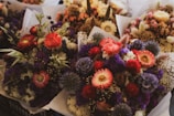 A collection of colorful flower bouquets featuring a variety of blossoms such as bright red and pink flowers, purple thistles, and delicate wildflowers. The arrangements are wrapped in white paper, creating an elegant presentation.