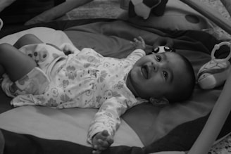 A baby is lying on a play mat, looking up with a joyful expression. The infant is wearing patterned clothing and is surrounded by soft toys. The environment is cozy and the baby appears to be relaxed and comfortable.