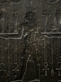 An ancient stone carving featuring a human figure in profile, surrounded by various symbolic objects. The figure is depicted with intricate detailing, including a traditional loincloth and distinctive hairstyle. Symbols such as ankh crosses and jars are positioned around the figure, suggesting a scene of ritual or offering.
