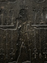 An ancient stone carving featuring a human figure in profile, surrounded by various symbolic objects. The figure is depicted with intricate detailing, including a traditional loincloth and distinctive hairstyle. Symbols such as ankh crosses and jars are positioned around the figure, suggesting a scene of ritual or offering.