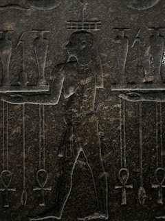 An ancient stone carving featuring a human figure in profile, surrounded by various symbolic objects. The figure is depicted with intricate detailing, including a traditional loincloth and distinctive hairstyle. Symbols such as ankh crosses and jars are positioned around the figure, suggesting a scene of ritual or offering.