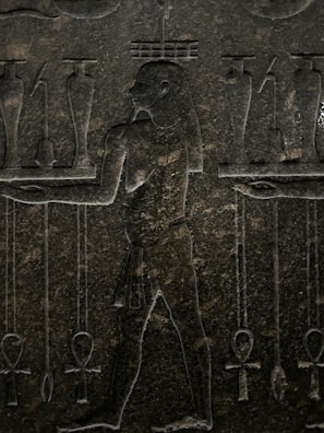 An ancient stone carving featuring a human figure in profile, surrounded by various symbolic objects. The figure is depicted with intricate detailing, including a traditional loincloth and distinctive hairstyle. Symbols such as ankh crosses and jars are positioned around the figure, suggesting a scene of ritual or offering.