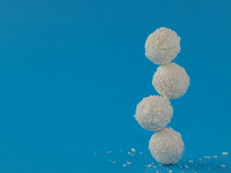 Four white, round confections coated with coconut flakes are stacked vertically against a solid blue background. Some coconut flakes are scattered around the base.