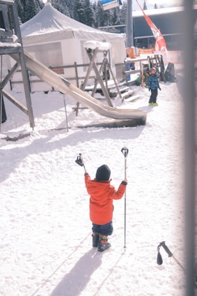 A child dressed in a bright red winter outfit stands in the snow holding ski poles, ready for skiing. Nearby, another child in a colorful jacket stands near a snow-covered playground structure, including a slide. In the background, there is a tent with snow on the roof, surrounded by other snowy equipment and a ski lift in the distance.
