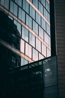 Glass façade of a modern building reflecting the sky and nearby structures. The windows are arranged in a grid pattern, with light and shadows creating an abstract geometric design.