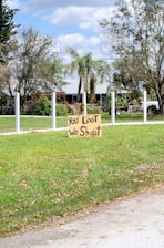 A handmade sign with the message 'You Loot, We Shoot' is placed on a patch of green grass in a suburban area. The background features a lightly colored house partially hidden behind trees and a clear sky with scattered clouds. A white fence outlines the property, and a person is visible near a parked vehicle.