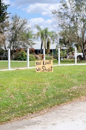 A handmade sign with the message 'You Loot, We Shoot' is placed on a patch of green grass in a suburban area. The background features a lightly colored house partially hidden behind trees and a clear sky with scattered clouds. A white fence outlines the property, and a person is visible near a parked vehicle.