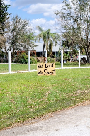 A handmade sign with the message 'You Loot, We Shoot' is placed on a patch of green grass in a suburban area. The background features a lightly colored house partially hidden behind trees and a clear sky with scattered clouds. A white fence outlines the property, and a person is visible near a parked vehicle.
