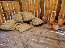 A rustic wooden interior with stacks of burlap sacks piled against a wooden wall. Next to the sacks are several clay pots with lids. The wooden floor and wall add a warm, natural texture to the setting.