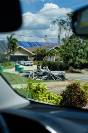 A residential street scene with a beige house in the background that has blue tarps on its roof. In front of the house, there is a pile of metal debris on the grass near the sidewalk. Green garbage bins are visible on the driveway, and there are palm trees and other greenery surrounding the area. The photo is taken from inside a vehicle, framed by parts of the dashboard.