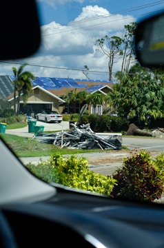 A residential street scene with a beige house in the background that has blue tarps on its roof. In front of the house, there is a pile of metal debris on the grass near the sidewalk. Green garbage bins are visible on the driveway, and there are palm trees and other greenery surrounding the area. The photo is taken from inside a vehicle, framed by parts of the dashboard.