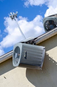An air conditioning unit is mounted on the exterior wall of a building. Above it, a telecommunications tower reaches into the sky, surrounded by fluffy white clouds under a bright blue sky. The structure of the building is made of light-colored concrete and the air conditioning unit is metallic with visible fans and grills.