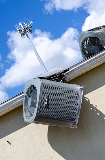 An air conditioning unit is mounted on the exterior wall of a building. Above it, a telecommunications tower reaches into the sky, surrounded by fluffy white clouds under a bright blue sky. The structure of the building is made of light-colored concrete and the air conditioning unit is metallic with visible fans and grills.