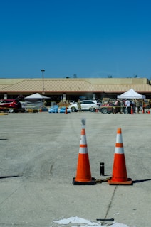 Two orange traffic cones are placed on a wide concrete area with a building in the background. The building has a tan-colored roof and there are several white tents set up outside. A few vehicles, including a white SUV and a red car, are parked nearby. People are gathered around the tents, and there are stacks of boxed goods, including bottled water, visible.