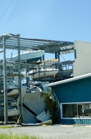A partially collapsed metal structure containing several boats stacked on different levels, with some sections of the building visibly damaged. The scene includes a blue building with a window that has 'Apparel Accessories' written on it. There is a palm tree next to the building, and the ground appears to be covered with debris.