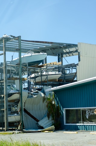 A partially collapsed metal structure containing several boats stacked on different levels, with some sections of the building visibly damaged. The scene includes a blue building with a window that has 'Apparel Accessories' written on it. There is a palm tree next to the building, and the ground appears to be covered with debris.